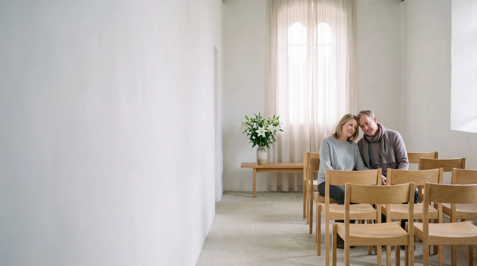 A couple sit close together, heads touching, in a small white chapel with pale oak chairs and white lilies