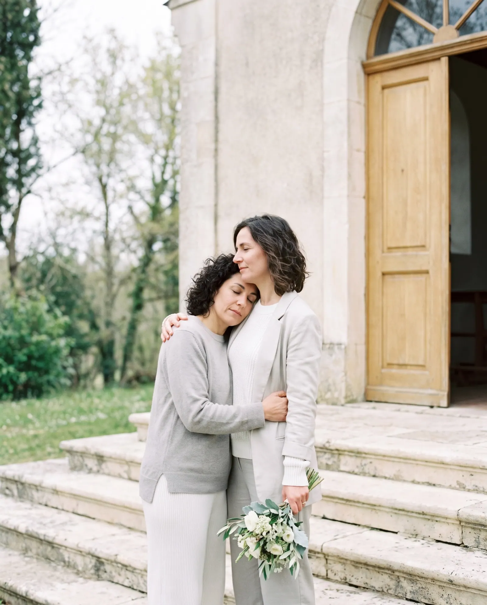Two sisters embrace on the pale stone steps of a small chapel, one holding white flowers