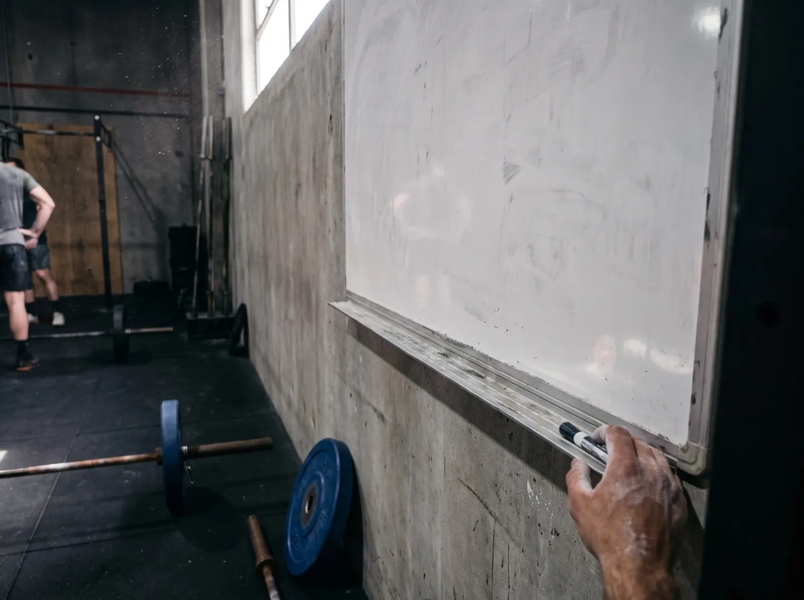 A chalk-dusted hand reaches for a marker on the ledge of a blank gym whiteboard, a barbell and a blue bumper plate on the floor
