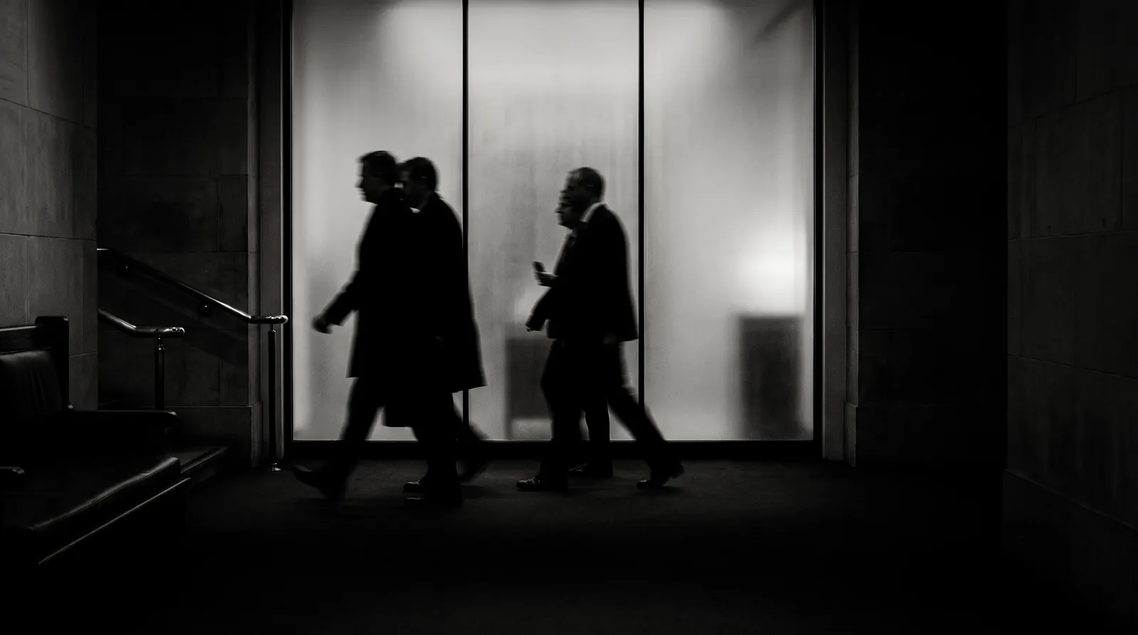 Black and white, long exposure: people in dark coats pass a lit frosted-glass wall as blurred silhouettes