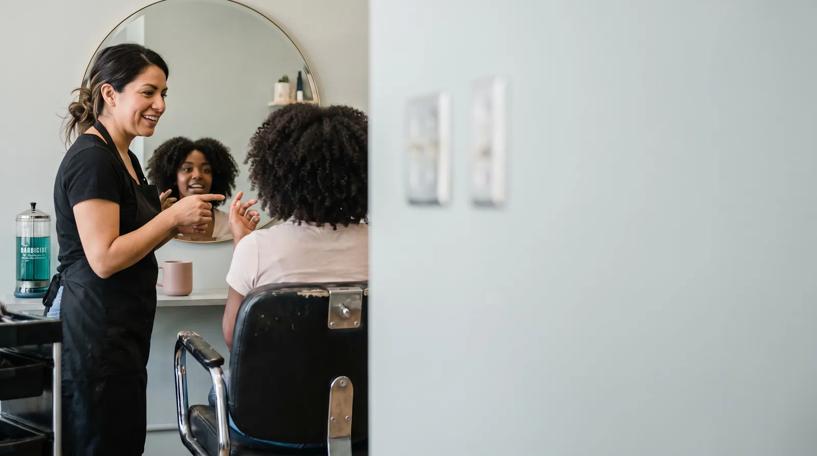 A stylist in a black apron smiles at a client with curly hair in a round salon mirror while talking through a new cut