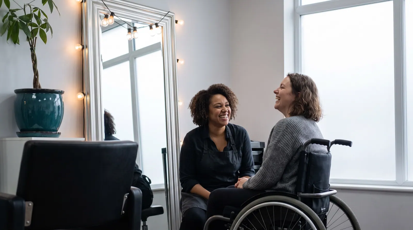 A stylist talks at eye level with a laughing client who uses a wheelchair at a styling station