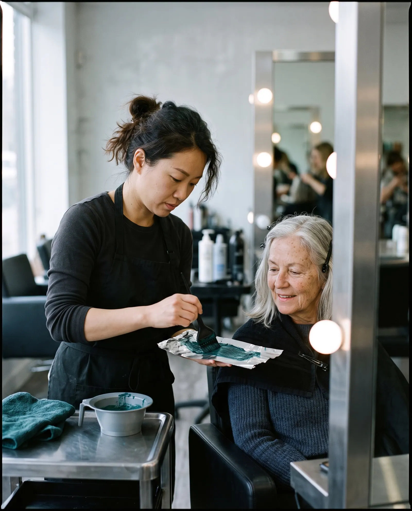 A colourist brushes dye onto a foil beside a smiling older client with long grey hair