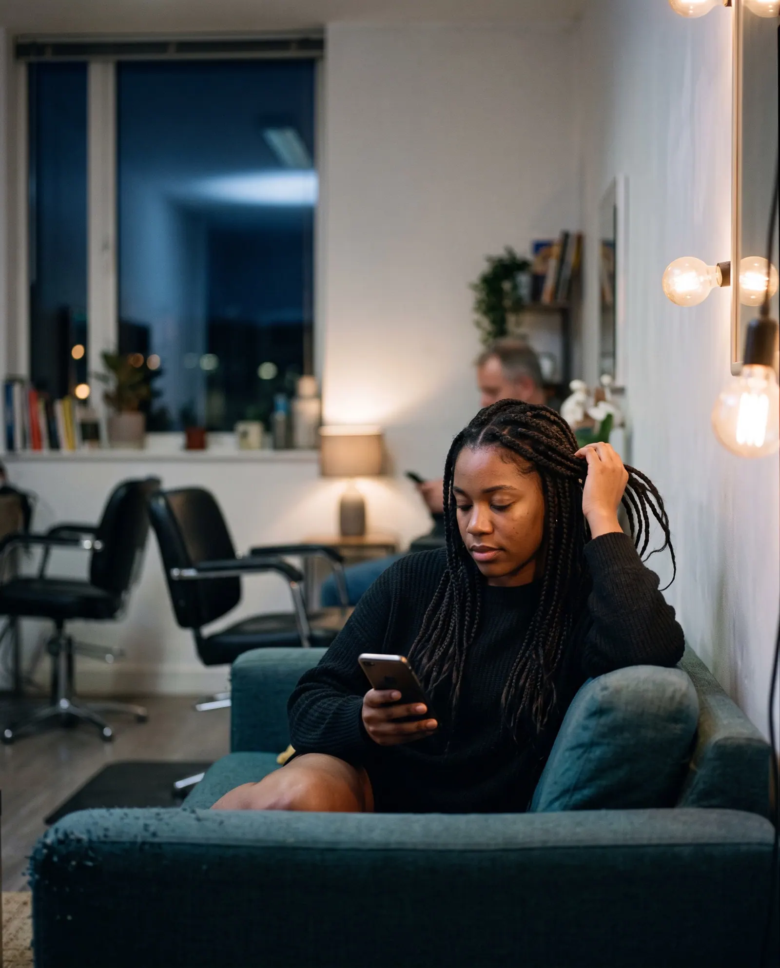 A young woman with long braids looks at her phone on a sofa, a hand in her hair