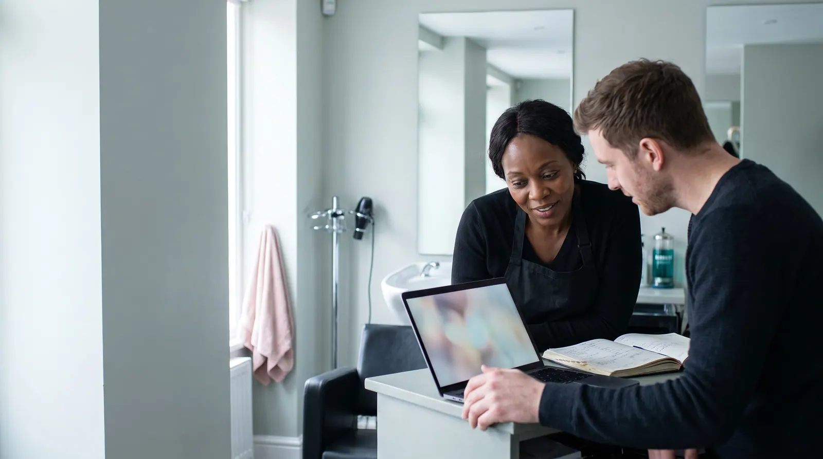 A web agency owner shows a laptop to a salon owner in a black apron at a reception desk beside an open appointment book